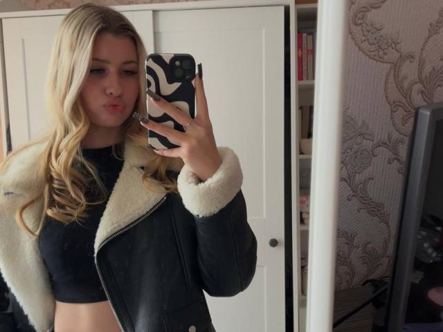 EmilyMorlino with long blonde hair, pouty lips, wearing a black leather jacket with white fur, black top, holding a black and white phone, mirror selfie.