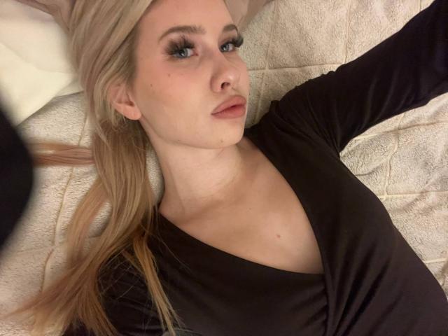 EmilyMorlino with blonde hair, blue eyes, and full lips, lying down, wearing a black V-neck shirt, showing a slight smile, with a relaxed and slightly seductive look.