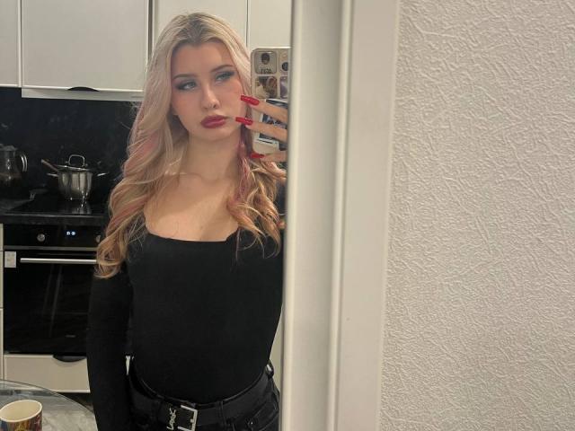 EmilyMorlino with long blonde hair, red lipstick, and black off-shoulder top, holding phone, wearing black pants, mirror selfie, white textured wall, red nails, kitchen background.