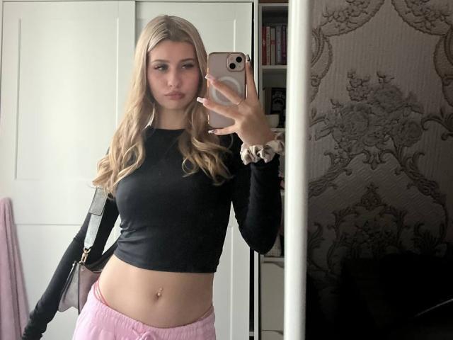 EmilyMorlino with blonde hair, black crop top, pink pants, holding phone in mirror, showing her tits and navel piercing, in a mirror selfie. Her ass is not visible.