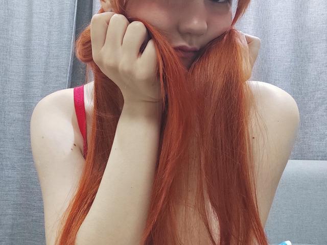 SheritaChieng with long red hair, hands holding her hair, pink bra straps, pale skin, slight smile, looking down, gray curtain in background, soft lighting, intimate pose.