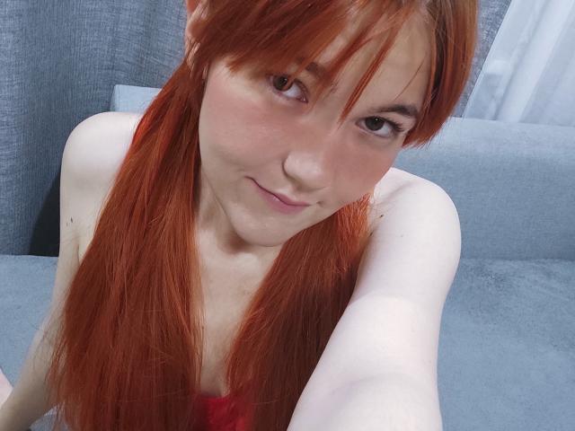 SheritaChieng, com maria-chiquinhas vermelhas, pele clara e um leve sorriso, exibindo seus ombros nus e um toque de lingerie vermelha, tirando uma selfie com o braço direito..