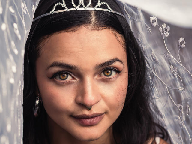 LucyySky, con ojos marrones, cabello negro y una tiara de diamantes, lleva un velo transparente con motivos florales y un piercing en la nariz, sonriendo suavemente a la cámara..