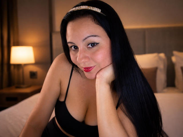 Gabriela Orozco, met zwart haar, draagt een hoofdband, een zwarte beha en rode lippenstift, met haar hoofd op haar hand rustend, haar borsten zichtbaar, zittend op een bed, kijkend naar de camera..