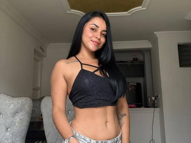 HikariChloe with long black hair, wearing a sparkly black crop top, exposing her small tits and tattoos, standing in a room, smiling with hands in silver pants.