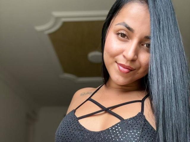 HikariChloe has long black hair, brown eyes, and a subtle smile. She's wearing a black strappy top, showing her tits. Tattoo on her left shoulder.