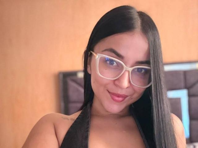 HikariChloe mit langem, schwarzem Haar, blauen Augen, die klare Brille trägt, lächelt mit rotem Lippenstift, zeigt ein kleines Tattoo an der linken Schulter, trägt ein schwarzes Top mit Halbschalen, in einem Schlafzimmer..