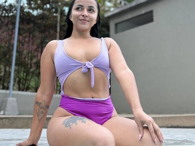 HikariChloe, uma latina com a pele úmida, vestindo um top de biquíni roxo amarrado e calcinha de cintura alta combinando, sentada em uma piscina, mostrando tatuagens no braço esquerdo e na coxa direita..