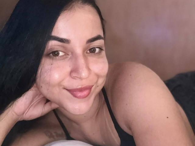 A HikariChloe tem cabelo escuro, olhos castanhos e um sorriso adorável. Ela está deitada, vestindo uma blusa preta, com a mão embaixo do queixo. Seus seios estão parcialmente visíveis..