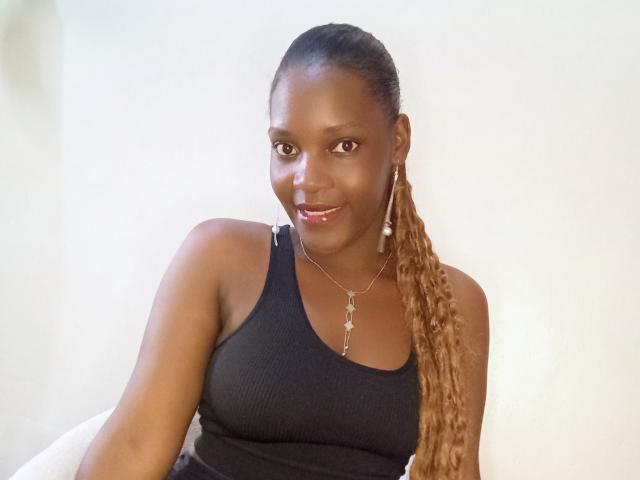 NaxCum a la peau brun foncé, de longs cheveux bruns bouclés, de petites seins et porte un débardeur noir à rayures. Elle a un léger sourire. Elle porte un collier et des boucles d'oreilles..