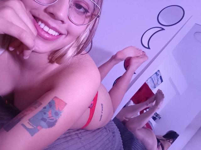RubyLane with blonde hair, glasses, and red panties, laying on a bed, smiling, showing a tattoo on her arm, and her ass, with another person's feet in the background.