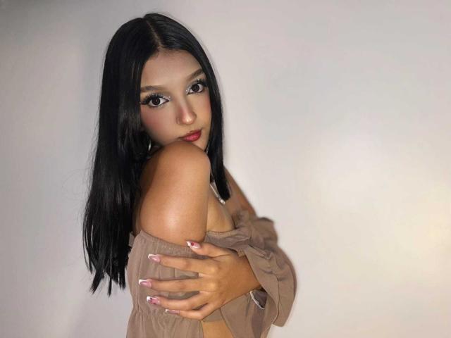 PaulineRey with long black hair, brown off-shoulder top, and pink nails, looking over shoulder with hands on chest, showing bare shoulder and soft expression.