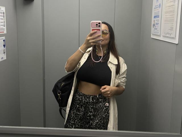 GalaMalkova com seios médios, top cropped preto, casaco branco, calças com estampa de leopardo, segurando um celular rosa, selfie no espelho, usando óculos, bolsa preta, dentro de um elevador, cabelo castanho longo..