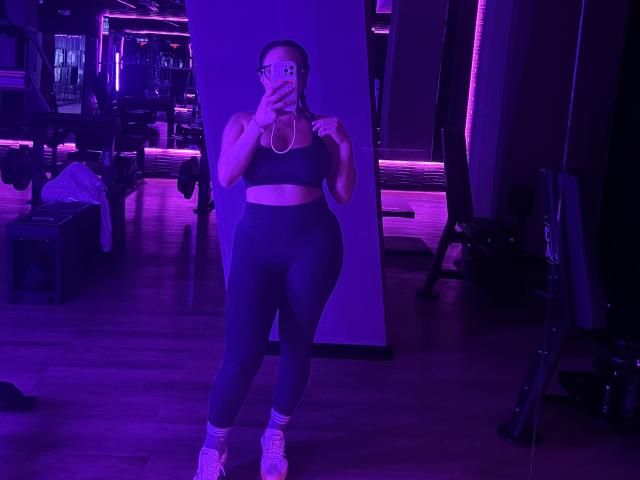 GalaMalkova stands in a gym, wearing a black sports bra and high-waisted leggings, holding a phone, with purple lighting, showing her big ass and small tits.