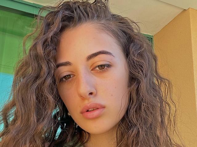 BokSinha has wavy brown hair, green eyes, and full pink lips with a slight pout. Her fair skin is slightly flushed, and she looks directly at the camera.
