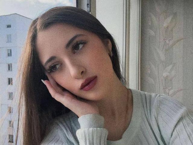 BokSinha with long brown hair, subtle makeup, and pink lipstick, resting her hand on her cheek, wearing a gray rib-knit sweater, looking to the side with a gentle expression.