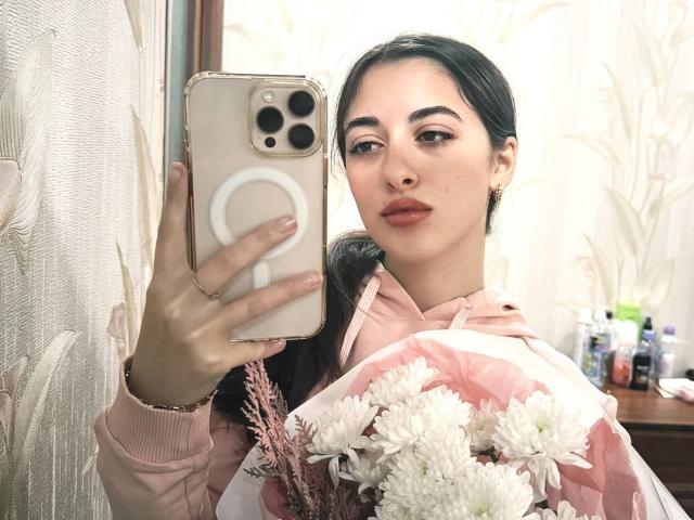 BokSinha with dark hair in a ponytail, pink hoodie, holding white flowers, taking a selfie with a gold iPhone, pink nails, and subtle makeup.