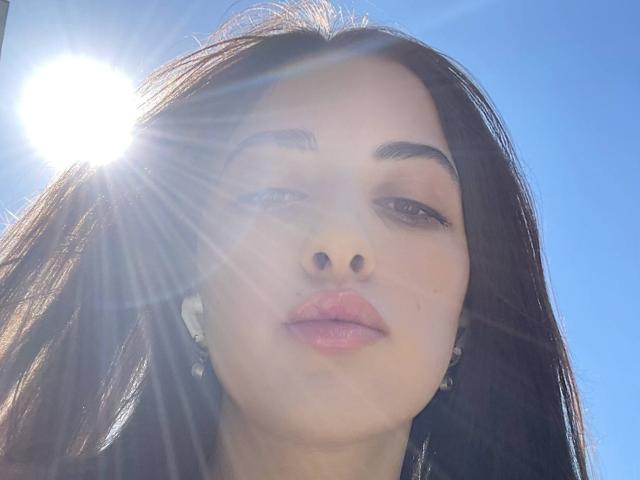 BokSinha's face with sun flare, dark straight hair, brown eyes, pink lips, small nose, wearing white earbuds, blue sky background, sun to the left, light rays in front.