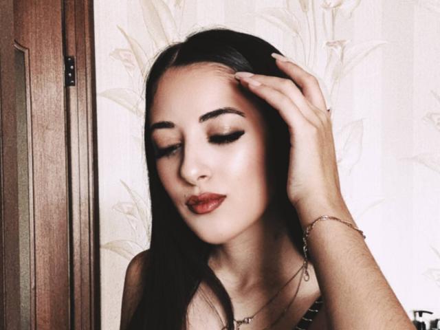 BokSinha has long black hair, dark eyeliner, and red lipstick, with a hand in her hair, wearing a bracelet, and a necklace, looking down with a subtle smile.