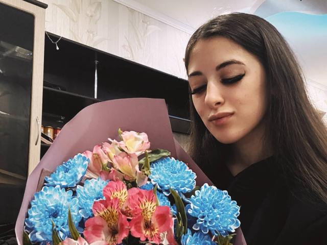 BokSinha with long brown hair, black shirt, holding colorful bouquet of pink, blue, and red flowers, looking down with subtle smile.