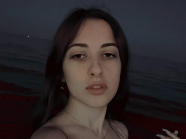 BokSinha with pale skin, dark brown hair, and small tits, has a neutral expression, slightly parted lips, and dark eyebrows in a dimly lit, dark-colored photo.