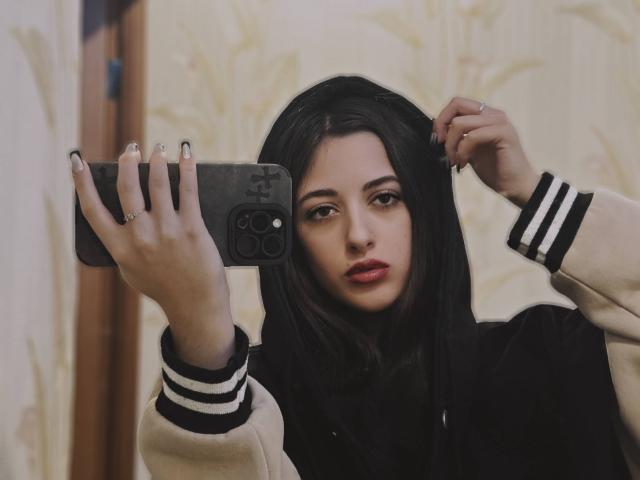 BokSinha with dark hair, holding a phone, taking a selfie, wearing a black and beige jacket with white stripes, red lips, and a ring on her finger.