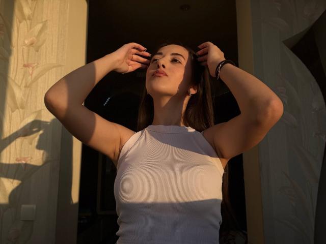 BokSinha with arms raised, hands in dark brown hair, wearing a white sleeveless top, sunlight on her face and chest, dark background, bracelet on right wrist, confident expression.