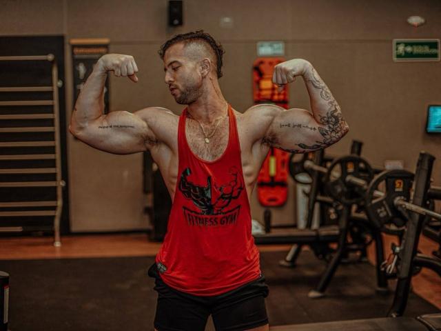 AllanDaf, gespierd, spieren aanspannend, rode tanktop, zwarte korte broek, tatoeages op de armen, geschoren zijkanten, baard, ketting, sportschool, biceps, borharen, geconcentreerd, staand, gewichten op de achtergrond, bezweet..