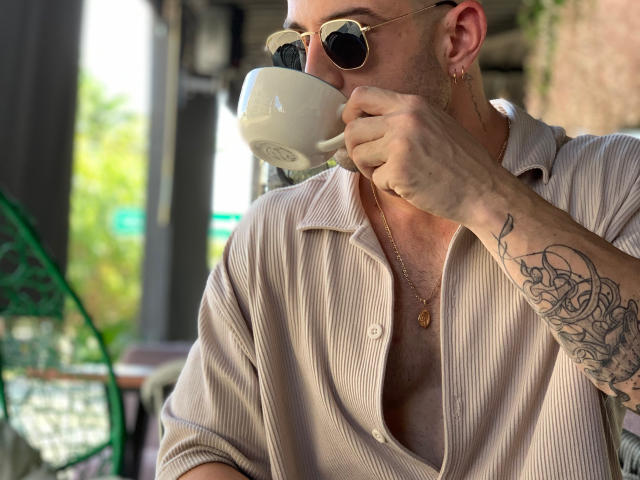 AllanDaf mit einer weißen Tasse, der ein beiges Rippshirt, eine goldene Halskette und eine Sonnenbrille trägt, trinkt etwas, zeigt einen tätowierten Arm und trägt goldene Ohrringe, draußen an einem sonnigen Tag..
