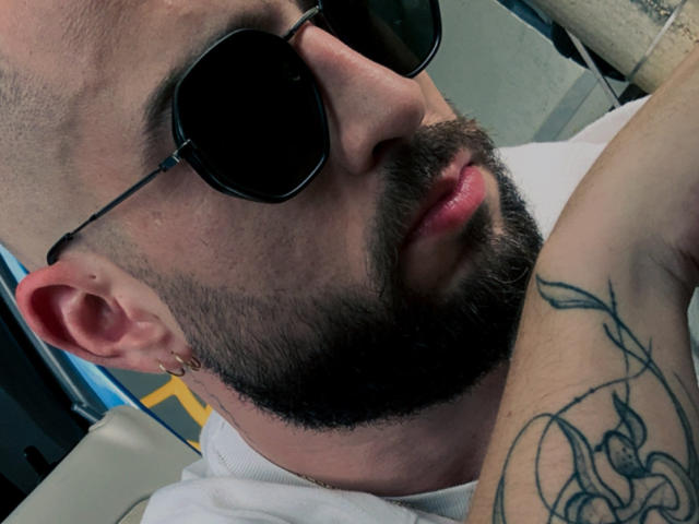 Alannedaf, com barba, usando óculos de sol escuros, uma camisa branca e brincos de ouro, mostrando uma tatuagem no braço, com lábios rosados levemente entreabertos..