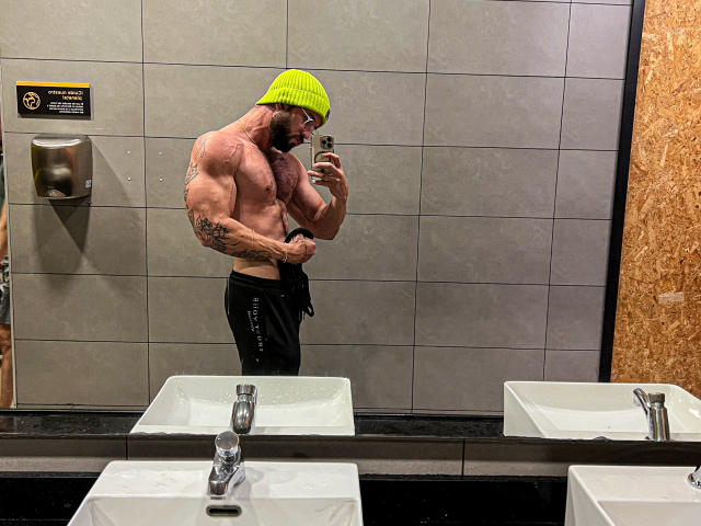 Gespierde AllanDaf met tatoeages, een neon groene muts, zwarte broek, die een spiegel-selfie maakt in een badkamer. Zijn grote, goed gedefinieerde billen zijn zichtbaar. Hij heeft een volle baard en houdt een telefoon vast..