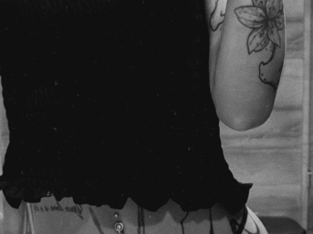 A blusa preta de SensualMila, o braço tatuado com uma grande flor e a piercing no umbigo visível. Foto em preto e branco, com textura, da parte superior do corpo..