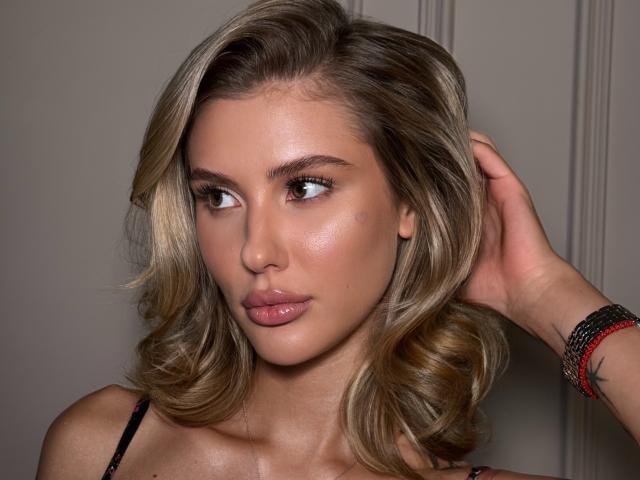 SensualMila has blonde, wavy hair, light skin, and full pink lips. She's looking to the side with a hand in her hair, wearing a black strap.