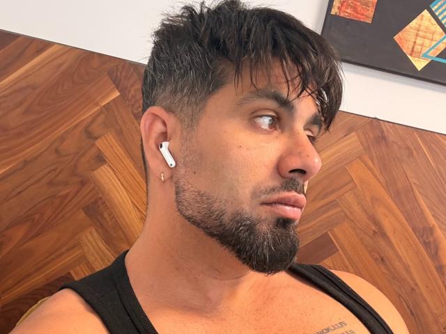 Gasston69 has short, dark, messy hair, a beard, black tank top, white earbuds in, and a tattoo on his left chest. His eyes are looking to the side.