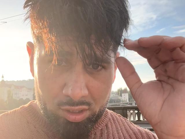 Gasston69 has a medium skin tone, dark messy hair, a beard, and a nose ring, wearing a pinkish sweater, with a sunlit hand making a peace sign near his face.