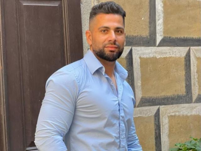 Gasston69, a muscular dude with a trimmed beard and short brown hair, wearing a tight light blue shirt, showing his toned chest and arms.