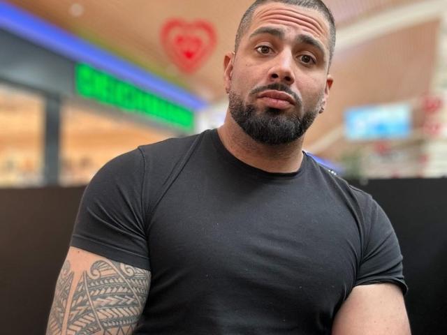 Gasston69, a muscular dude with a black beard and tattoos on his right arm, wearing a tight black shirt, looking surprised with a slightly raised eyebrow.