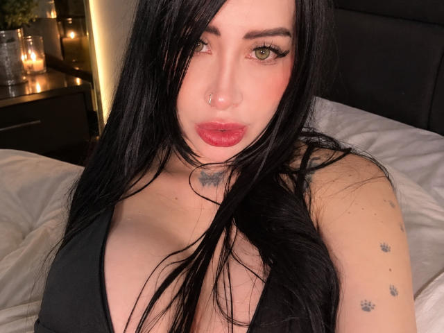Kkole has long black hair, green eyes, and red lipstick. She's wearing a black top, showing her tits, with tattoos on her neck and shoulder. She's looking at the camera.