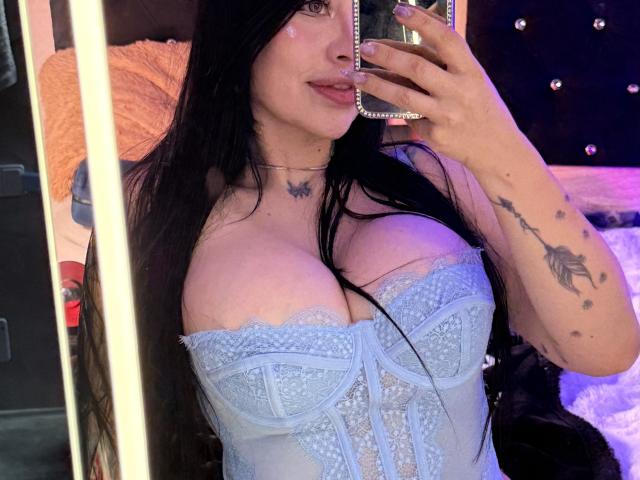 Kkole with large tits in a blue lace corset, taking a mirror selfie with long black hair, a butterfly tattoo, and a scorpion tattoo on her right arm.