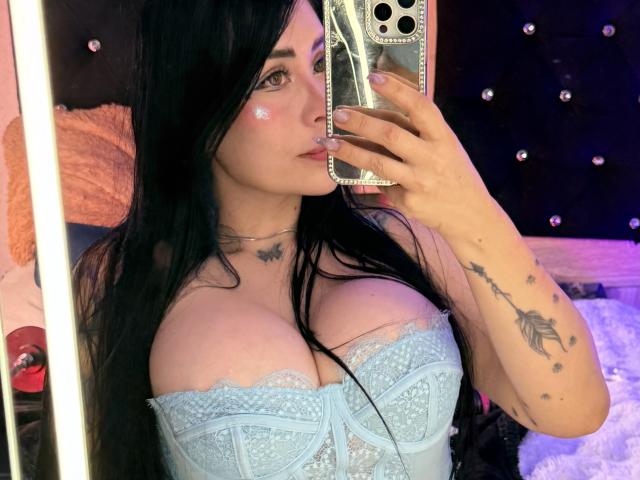 Kkole with big tits, wearing a blue lace top, black hair, holding a phone, glitter on cheek, tattoo on arm, taking a mirror selfie, in a dimly lit room.