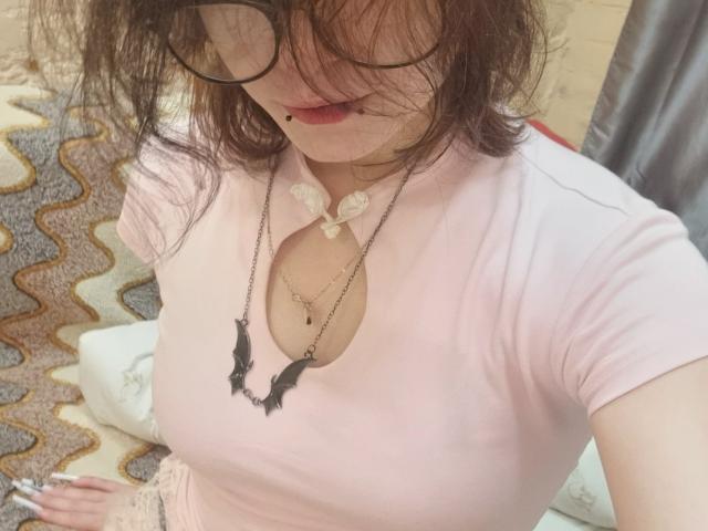 SophiaRenlay with messy brown hair, glasses, pink shirt, black bat necklace, small cleavage, and a cute smile, looking down at herself, holding a cigarette.