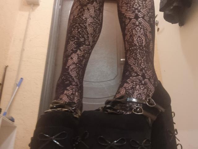 SophiaRenlay's legs in black lace stockings, black high-heeled boots with straps, standing in a doorway. Visible door, white wall, and blue-handled broom in background.