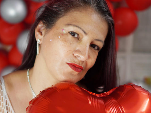 MilenneSexy houdt rode, hartvormige ballonnen vast, heeft rode lippenstift, glinsterende oogmake-up en een schattig gezicht, en draagt een parelketting en oorbellen. Haar donkere haar is recht..