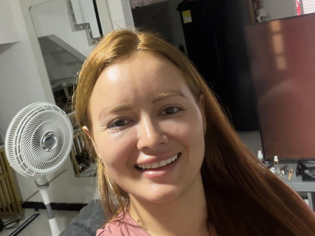 MiaLuxury, avec ses cheveux roux, souriante, peau claire, petit nez et lèvres pulpeuses, se tient devant un ventilateur blanc, vêtue d'une chemise rose, dans une pièce bien éclairée..