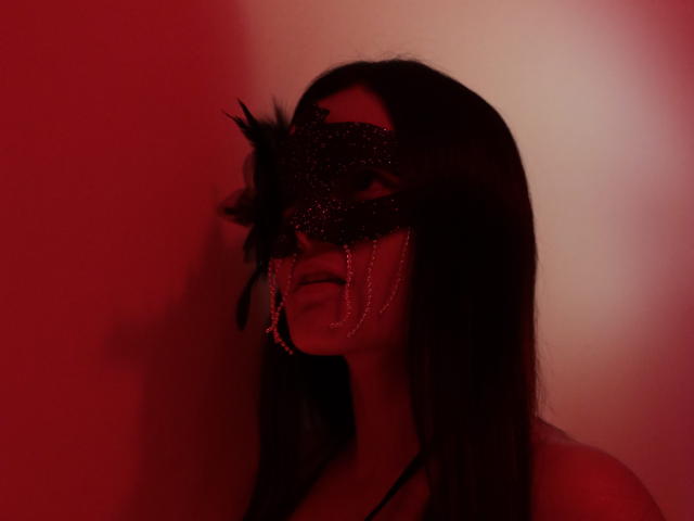 MiaLuxury with long dark hair, wearing a glittery black mask with feathers and chains, red-lit face, slightly open mouth, shadow on wall, subtle sensual expression.