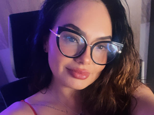 GalaMalkova with large, black-rimmed glasses, wavy brown hair, and glossy pink lips, wearing a red spaghetti strap top, smiling softly. Her tits are partially visible.