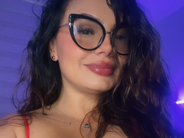 GalaMalkova with glossy pink lips, wavy brown hair, black cat-eye glasses, and red strap dress, showing a subtle smile. She's got cute ear piercings and a small necklace.