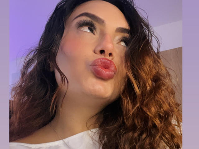 GalaMalkova has curly brown hair, pouty pink lips, and raised eyebrows, looking to the side with a playful expression, wearing a white shirt.