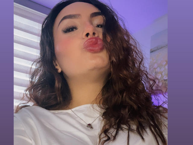 GalaMalkova with wavy brown hair, pouty pink lips, and thick eyebrows, wearing a white shirt, necklace with small pendant, and black ear studs, in a purple-lit room.