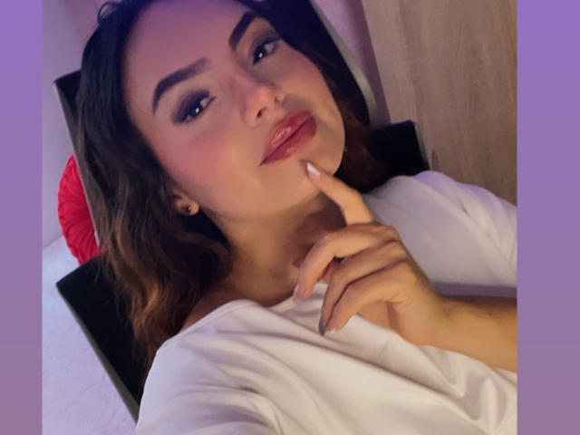 GalaMalkova with dark brown hair, makeup, and pink lipstick, resting her chin on her finger, wearing a white top, looking at the camera with a subtle smile.